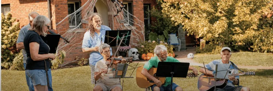 Musicians with a fiddle and acoustic guitars playing from music stands on a front lawn decorated for fall